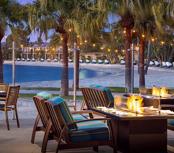 Scenic beachfront dining at Dockside 1953 with cozy fire pit tables, string lights, and a view of Mission Bay at dusk steps away from Bahia Beach and Boat Club.